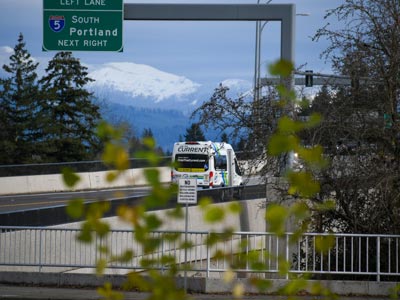 A C-TRAN vehicle for The Current on-demand service is spotted through the trees from the Salmon Creek Park and Ride. In the distance, there are snow-capped mountains, and tall evergreen trees.