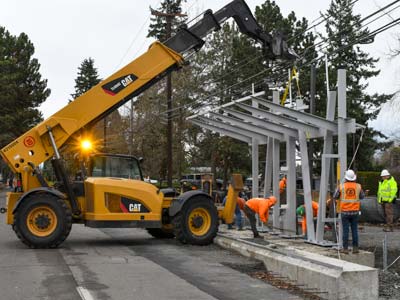 A construction crew places a steel shelter at a Vine station that is under construction. There is a crane in the street lifting the shelter onto the platform.