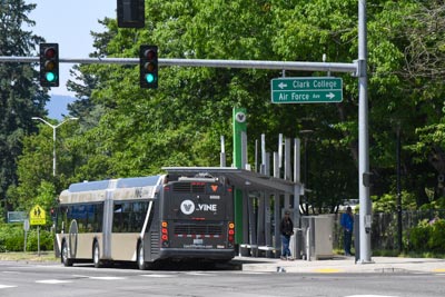 A C-TRAN Vine vehicle sits next to a station on Fort Vancouver Way at Air Force Ave between Clark College and the VA. Passengers stand at the station. Large, green trees line the street.